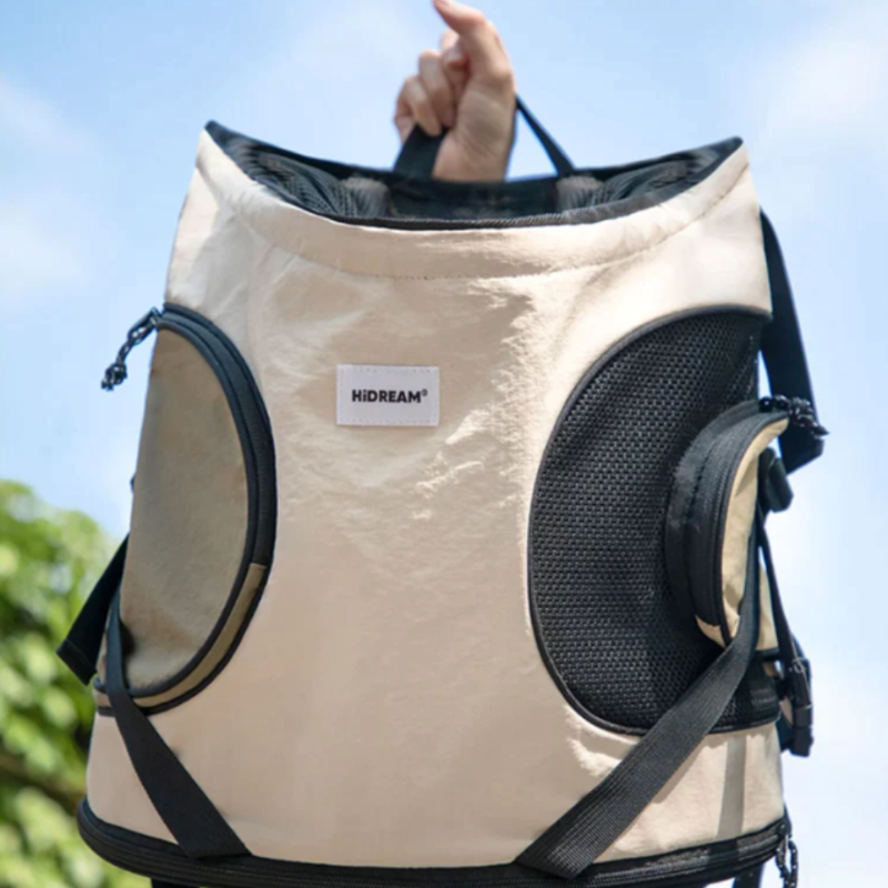 A beige and black pet backpack carrier with mesh windows and adjustable straps, being held by a person.